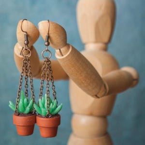 May include: Dangle earrings with miniature succulent plants in terracotta pots. The earrings have bronze-coloured chains and hooks. The background is a soft blue, and the earrings are held by a wooden mannequin.