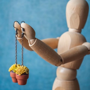 Op de afbeelding: Een houten mannequin houdt een paar oorbellen vast. Elke oorbel heeft een kleine terracotta pot gevuld met gele en groene vetplanten. De oorbellen hangen aan donkere metalen kettingen en haken tegen een blauwe achtergrond.