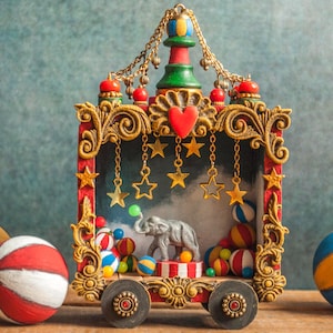 Op de afbeelding: Een decoratieve circus-thema display met een miniatuur olifant, kleurrijke ballen en een rood en goudkleurig sierlijk frame. De display bevat hangende sterornamenten en wordt geflankeerd door drie geschilderde ballen.