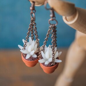 May include: Earrings with miniature succulent plants in terracotta pots. The white succulent flowers hang from copper-coloured chains and hooks. The earrings are displayed against a blue backdrop.