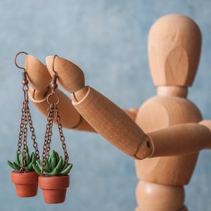 Puede incluir: Pendientes con pequeñas suculentas en macetas de terracota, suspendidas de cadenas color cobre. Los pendientes son sostenidos por un maniquí de madera sobre un fondo azul claro. Un accesorio único.