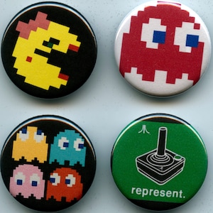 May include: Four round pinback buttons with pixelated designs. One features a yellow Pac-Man character, another a red ghost, and a third shows four colorful ghosts. The last button has a green background with a black joystick and the word "represent."