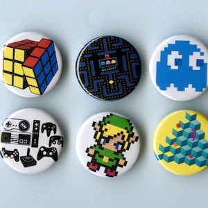 May include: A collection of ten round pinback buttons featuring retro video game designs. The buttons showcase iconic graphics from games like Pac-Man, Donkey Kong, and Atari, along with a Rubik's Cube and a pixelated Link.