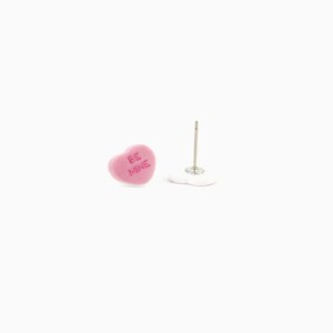May include: Pink heart-shaped stud earrings with the text "BE MINE" in a light pink color. The earrings are on a white background. One earring is upright, and the other is lying down.