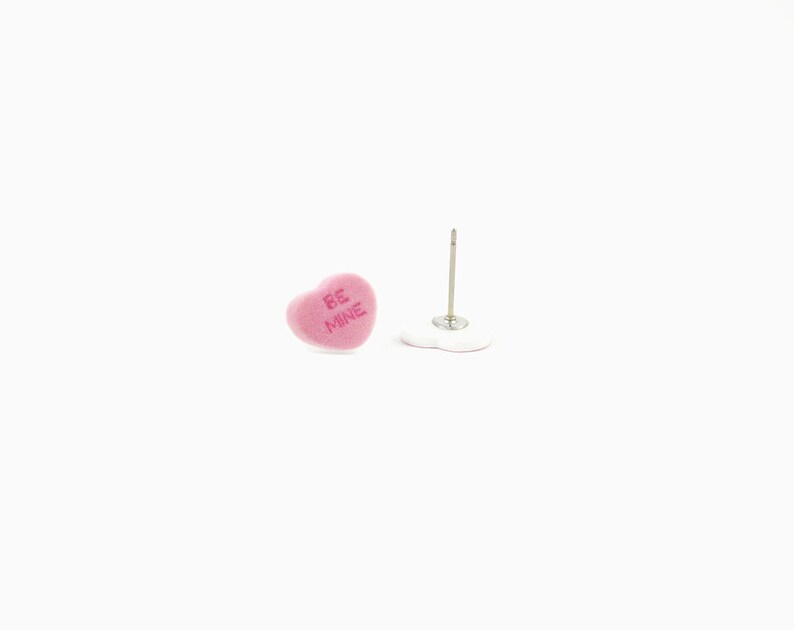 May include: Pink heart-shaped stud earrings with the text "BE MINE" in a light pink color. The earrings are on a white background. One earring is upright, and the other is lying down.