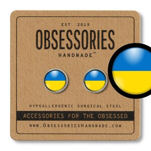Puede incluir: Un par de pendientes con la bandera de Ucrania. Los pendientes están hechos de acero quirúrgico hipoalergénico y son vendidos por la empresa Obsessories Handmade. El texto en la tarjeta dice "EST. 2015 OBSESSORIES HANDMADE TM HYPOALLERGENIC SURGICAL STEEL ACCESSORIES FOR THE OBSESSED WWW.OBSESSORIESHANDMADE.COM"