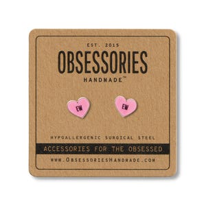 May include: Pink heart-shaped earrings with the word "EW" in black, presented on a brown card. The card features the text "OBSESSORIES HANDMADE" and "ACCESSORIES FOR THE OBSESSED".