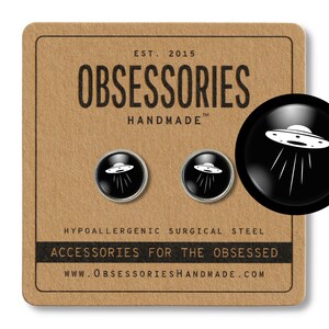 May include: A pair of black and silver cufflinks featuring a white UFO design. The cufflinks are displayed on a brown card with the text "OBSESSORIES HANDMADE" and "ACCESSORIES FOR THE OBSESSED".