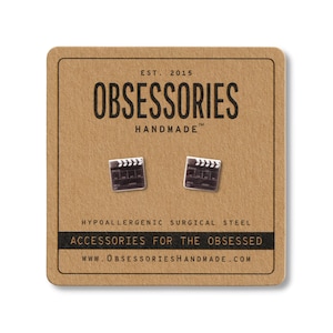 May include: Silver-coloured film clapperboard stud earrings displayed on a brown card. The card features the text "OBSESSORIES HANDMADE" and "ACCESSORIES FOR THE OBSESSED". The earrings are made of hypoallergenic surgical steel.