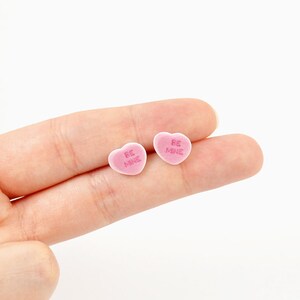May include: Two pink heart-shaped candies with the words "BE MINE" in pink lettering. The candies are small and appear to be made of a soft, sugary material. The candies are resting on a person's fingers.