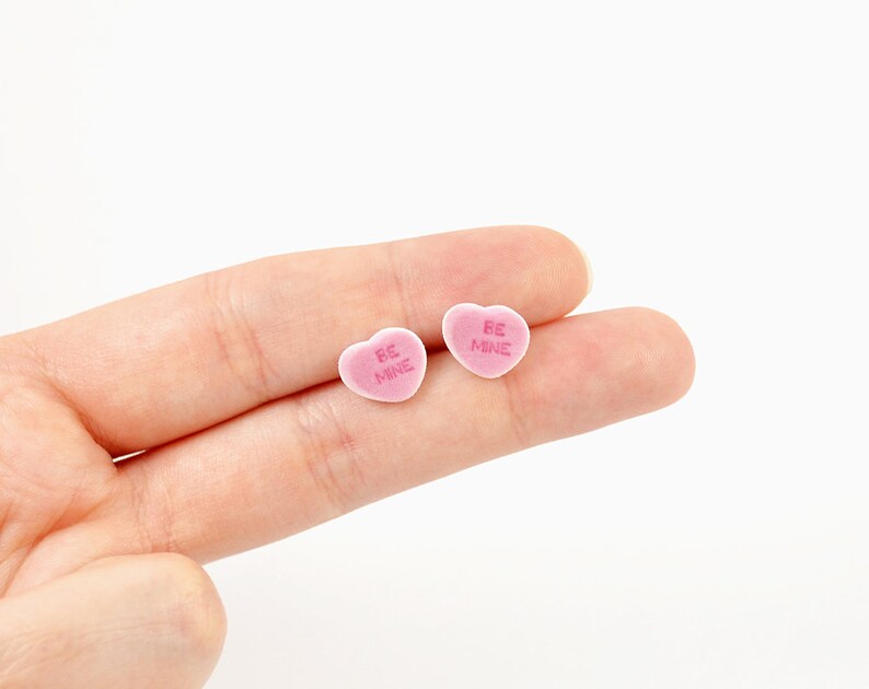 May include: Two pink heart-shaped candies with the words "BE MINE" in pink lettering. The candies are small and appear to be made of a soft, sugary material. The candies are resting on a person's fingers.