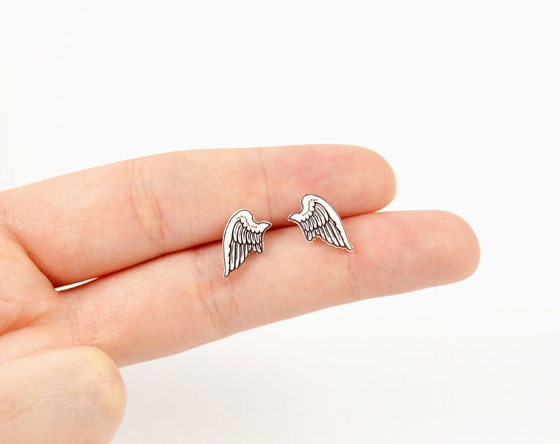 Puede incluir: Un par de pendientes peque&ntilde;os plateados con forma de alas. Cada pendiente presenta un detallado dise&ntilde;o de plumas y una forma ligeramente curvada, sugiriendo un dise&ntilde;o delicado. Los pendientes se muestran sobre un fondo neutro.