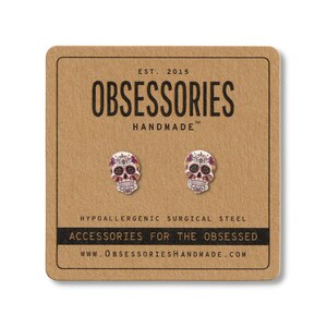 May include: A pair of skull-shaped stud earrings on a brown card. The earrings are silver with red and purple accents. The card reads "OBSESSORIES HANDMADE" and "ACCESSORIES FOR THE OBSESSED". The earrings are made of hypoallergenic surgical steel.
