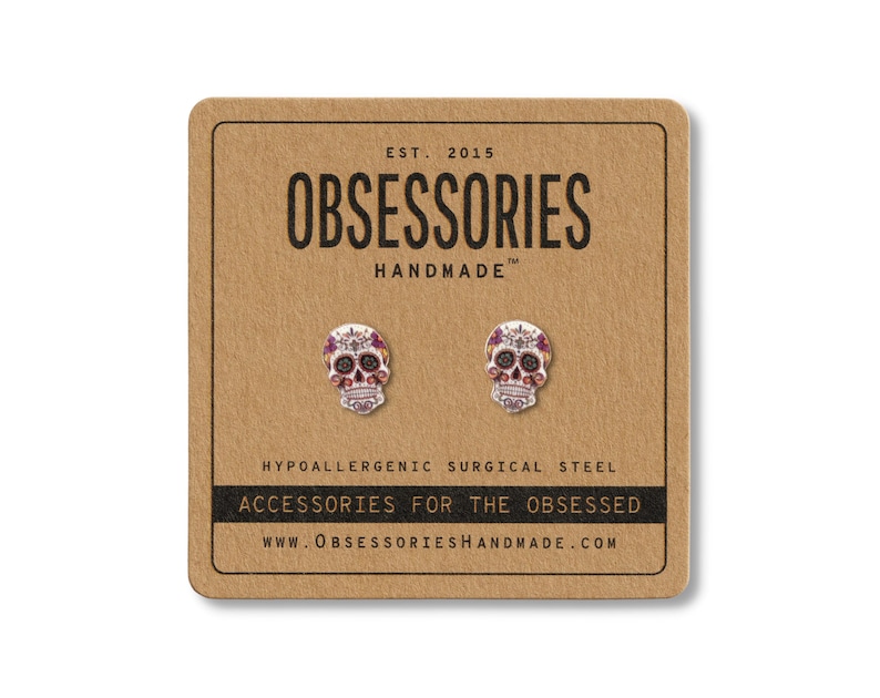 May include: A pair of skull-shaped stud earrings on a brown card. The earrings are silver with red and purple accents. The card reads "OBSESSORIES HANDMADE" and "ACCESSORIES FOR THE OBSESSED". The earrings are made of hypoallergenic surgical steel.