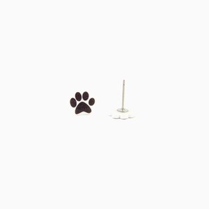 May include: A pair of stud earrings featuring a black paw print design. One earring is a complete paw print, while the other is a post with a white, flower-shaped backing. The earrings are set against a white background.
