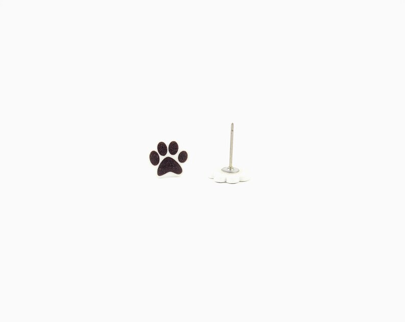 May include: A pair of stud earrings featuring a black paw print design. One earring is a complete paw print, while the other is a post with a white, flower-shaped backing. The earrings are set against a white background.