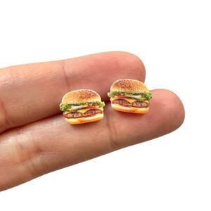 May include: Two miniature hamburger earrings. Each earring features a sesame seed bun, lettuce, tomato, cheese, and a burger patty. The earrings are small and detailed, perfect for a food-themed accessory.