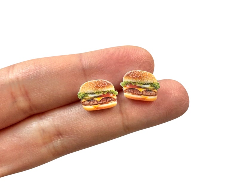 May include: Two miniature hamburger earrings. Each earring features a sesame seed bun, lettuce, tomato, cheese, and a burger patty. The earrings are small and detailed, perfect for a food-themed accessory.