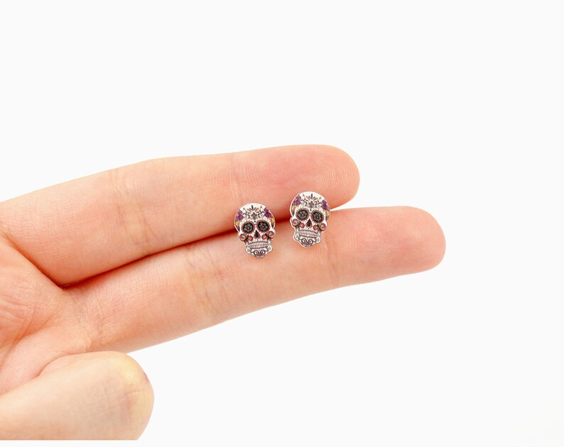 May include: A pair of small, silver-toned skull stud earrings. Each skull features intricate detailing with purple and black accents, including gem-like embellishments. The earrings are designed with a Day of the Dead theme.