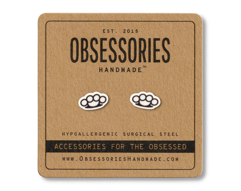 Puede incluir: Un par de pendientes de bot&oacute;n con forma de nudillos blancos y negros sobre una tarjeta marr&oacute;n. La tarjeta tiene el texto "OBSESSORIES HANDMADE" y "ACCESSORIES FOR THE OBSESSED". Los pendientes son de acero quir&uacute;rgico hipoalerg&eacute;nico.