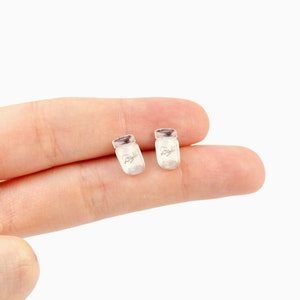 May include: Two small, light-colored earrings shaped like miniature mason jars. Each jar has a silver-colored lid and the word "Ball" printed on the front. The earrings are set against a plain white background.