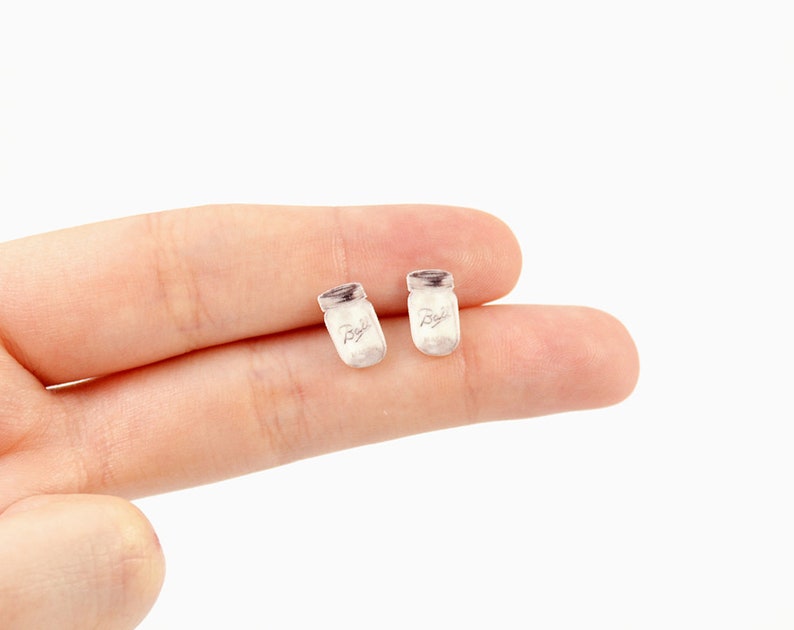May include: Two small, light-colored earrings shaped like miniature mason jars. Each jar has a silver-colored lid and the word "Ball" printed on the front. The earrings are set against a plain white background.