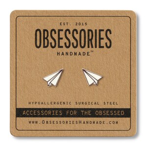 May include: A pair of paper airplane stud earrings on a brown card. The earrings are white with silver outlines. The card has the text "OBSESSORIES HANDMADE" and "ACCESSORIES FOR THE OBSESSED". The earrings are made of hypoallergenic surgical steel.