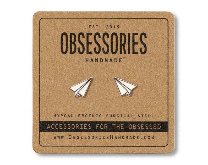May include: A pair of paper airplane stud earrings on a brown card. The earrings are white with silver outlines. The card has the text "OBSESSORIES HANDMADE" and "ACCESSORIES FOR THE OBSESSED". The earrings are made of hypoallergenic surgical steel.