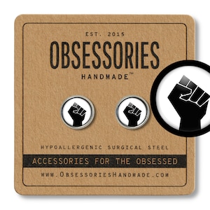 May include: A pair of black and white raised fist earrings made of hypoallergenic surgical steel. The earrings are on a brown background with the text "OBSESSORIES HANDMADE" and "ACCESSORIES FOR THE OBSESSED".