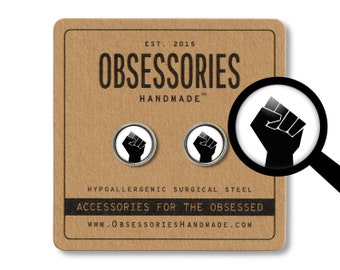 May include: A pair of black and white raised fist earrings made of hypoallergenic surgical steel. The earrings are on a brown background with the text "OBSESSORIES HANDMADE" and "ACCESSORIES FOR THE OBSESSED".