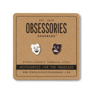 May include: A pair of hypoallergenic surgical steel stud earrings on a brown card. One earring is a white smiling mask, the other is a dark brown frowning mask. The card reads "OBSESSORIES HANDMADE" and "ACCESSORIES FOR THE OBSESSED".