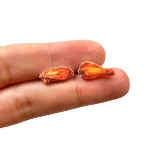Puede incluir: Se muestran dos alitas de pollo en miniatura, con una apariencia realista. Las alitas son de color marr&oacute;n dorado, con zonas m&aacute;s oscuras que sugieren una textura crujiente. La imagen est&aacute; sobre un fondo blanco.