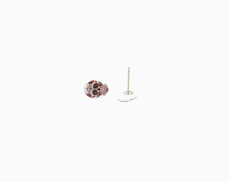 May include: A pair of Day of the Dead skull stud earrings. The earrings feature a colorful sugar skull design with intricate details in shades of purple, orange, and white. The post is silver-colored.