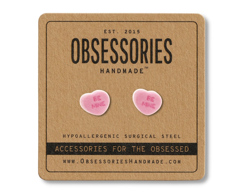May include: A pair of pink heart-shaped earrings with the text "BE MINE" on a brown card. The card reads "OBSESSORIES HANDMADE" and "ACCESSORIES FOR THE OBSESSED". The earrings are made of hypoallergenic surgical steel.