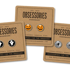 May include: Three sets of Halloween themed earrings on brown card stock. The first set features a pair of black and white ghost earrings. The second set features a pair of orange jack-o-lantern earrings. The third set features a pair of black and white witch earrings. Each card has the text "OBSESSORIES HANDMADE" and "EST. 2015".
