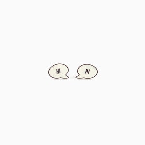 May include: A pair of cream-colored speech bubble earrings with the word "HI" in black lettering. The earrings have a black outline and are set against a white background.