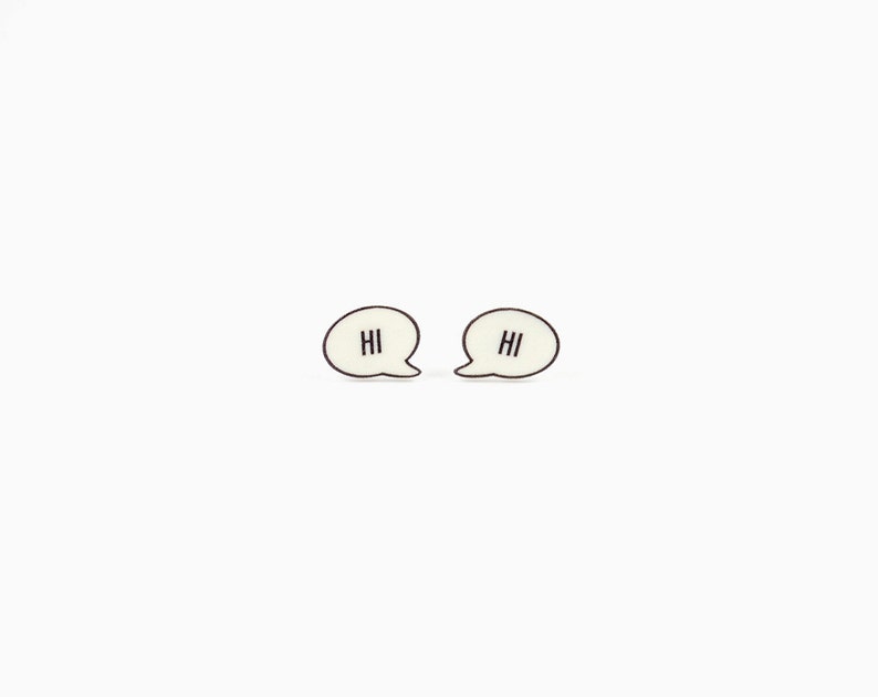 May include: A pair of cream-colored speech bubble earrings with the word "HI" in black lettering. The earrings have a black outline and are set against a white background.