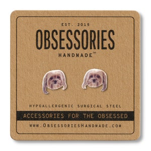 May include: A pair of dog-shaped earrings on a brown card. The card reads "OBSESSORIES HANDMADE" and "ACCESSORIES FOR THE OBSESSED." The earrings are hypoallergenic surgical steel.
