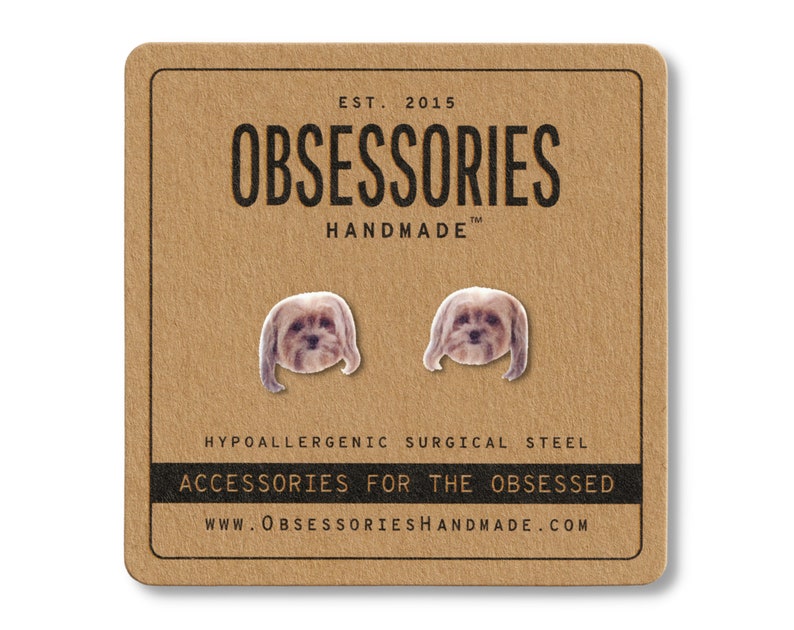 May include: A pair of dog-shaped earrings on a brown card. The card reads "OBSESSORIES HANDMADE" and "ACCESSORIES FOR THE OBSESSED." The earrings are hypoallergenic surgical steel.