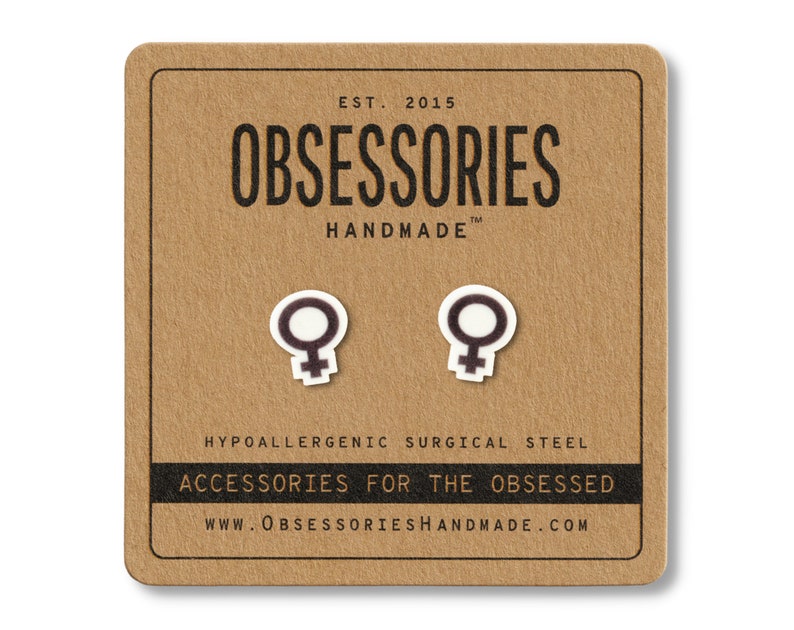 May include: A pair of white and black Venus symbol stud earrings on a brown card. The card reads "OBSESSORIES HANDMADE" and "ACCESSORIES FOR THE OBSESSED". The earrings are made of hypoallergenic surgical steel.