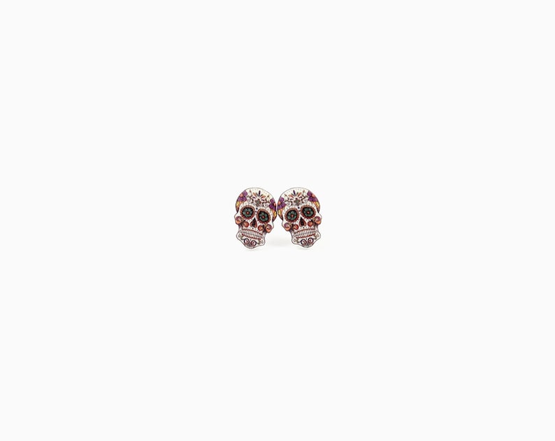 May include: A pair of skull-shaped stud earrings. The earrings feature a detailed design with a white base, adorned with purple, red, and black accents. The skull's eyes and other features are highlighted with small, colorful embellishments.
