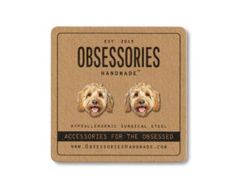 Puede incluir: Una tarjeta cuadrada, de color marrón claro, con el texto "OBSESSORIES HANDMADE" y dos pendientes con forma de cara de perro. La tarjeta también incluye el texto "ACCESSORIES FOR THE OBSESSED" y "HYPOALLERGENIC SURGICAL STEEL". También se incluye la dirección del sitio web.