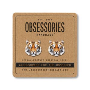 Pode incluir: Brincos de pressão com cabeças de tigre em um cartão marrom. Os brincos apresentam rostos de tigre detalhados com marcas laranja, pretas e brancas. O cartão diz "OBSESSORIES HANDMADE" e "ACCESSORIES FOR THE OBSESSED".