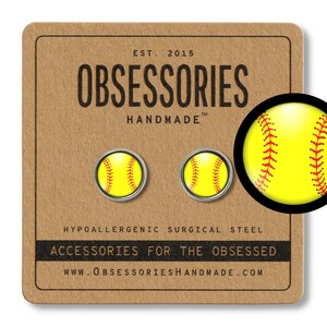 Pode incluir: Um par de brincos de aço cirúrgico hipoalergênico com uma bola de softball amarela com costuras vermelhas. Os brincos são exibidos em um cartão marrom com o texto "OBSESSORIES HANDMADE" e "ACCESSORIES FOR THE OBSESSED".