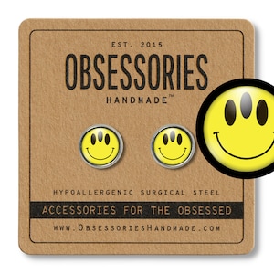 May include: A pair of yellow smiley face earrings with three eyes. The earrings are made of hypoallergenic surgical steel. The earrings are on a brown card with the text "OBSESSORIES HANDMADE" and "ACCESSORIES FOR THE OBSESSED".