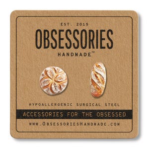 Op de afbeelding: Een bruine kaart met de tekst "EST. 2015 OBSESSORIES HANDMADE TM" en de tekst "HYPOALLERGENIC SURGICAL STEEL ACCESSORIES FOR THE OBSESSED WWW.OBSESSORIESHANDMADE.COM". De kaart heeft twee afbeeldingen van brood.