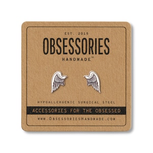 Puede incluir: Pendientes de bot&oacute;n plateados con forma de alas de &aacute;ngel sobre una tarjeta marr&oacute;n. La tarjeta tiene el texto "OBSESSORIES HANDMADE" y "ACCESSORIES FOR THE OBSESSED". Los pendientes son de acero quir&uacute;rgico hipoalerg&eacute;nico.
