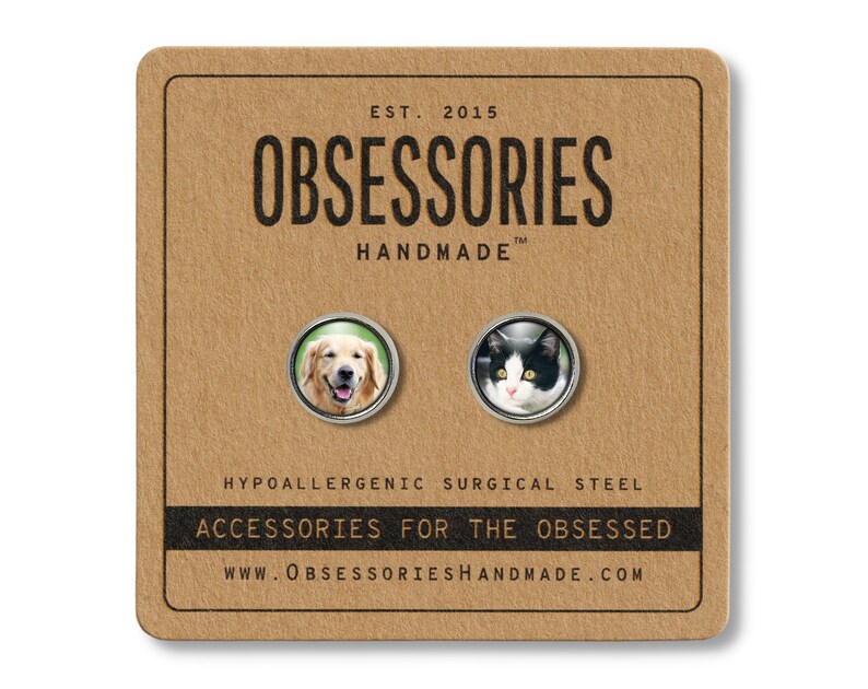 May include: A pair of hypoallergenic surgical steel earrings featuring a golden retriever and a black and white cat. The earrings are on a brown card with the text "OBSESSORIES HANDMADE" and "ACCESSORIES FOR THE OBSESSED".