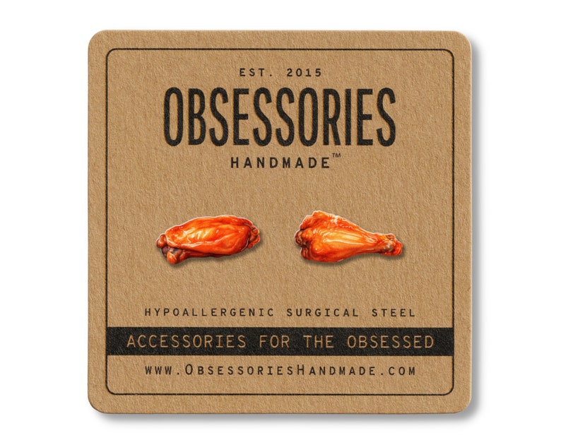 Puede incluir: Una tarjeta cuadrada marr&oacute;n con el texto "OBSESSORIES HANDMADE" y "ACCESSORIES FOR THE OBSESSED". Se presentan dos ilustraciones realistas de alitas de pollo. La tarjeta tambi&eacute;n indica "HYPOALLERGENIC SURGICAL STEEL."