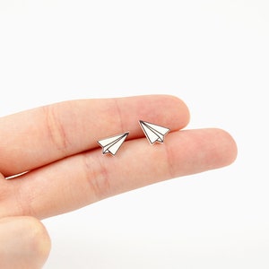 May include: Two small, white paper airplane earrings with black outlines. The earrings are displayed on a light background. The paper airplane design is a classic and whimsical motif.